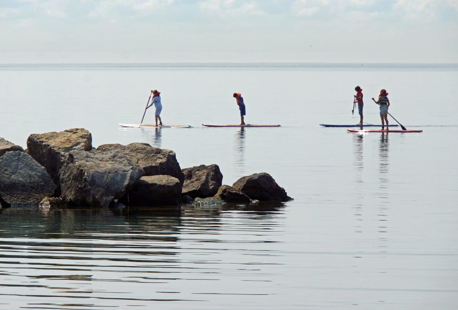 4 people paddle board on a still lake past a rocky shore.