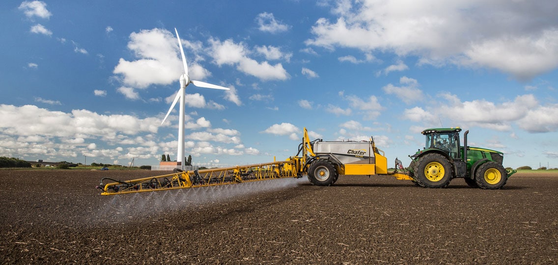 A large tractor drives along a flat field and sprays the crops. In the background a wind turbine sits against the cloudy sky.