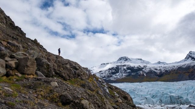 A person stands on a steep rocky cliff that overlooks a glacier. In the distance stand snow covered mountains.