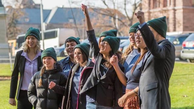 A group of young activists stand together and laugh. 2 raise their hands in victory. They all wear green toques.