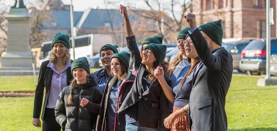 A group of young activists stand together and laugh. 2 raise their hands in victory. They all wear green toques.