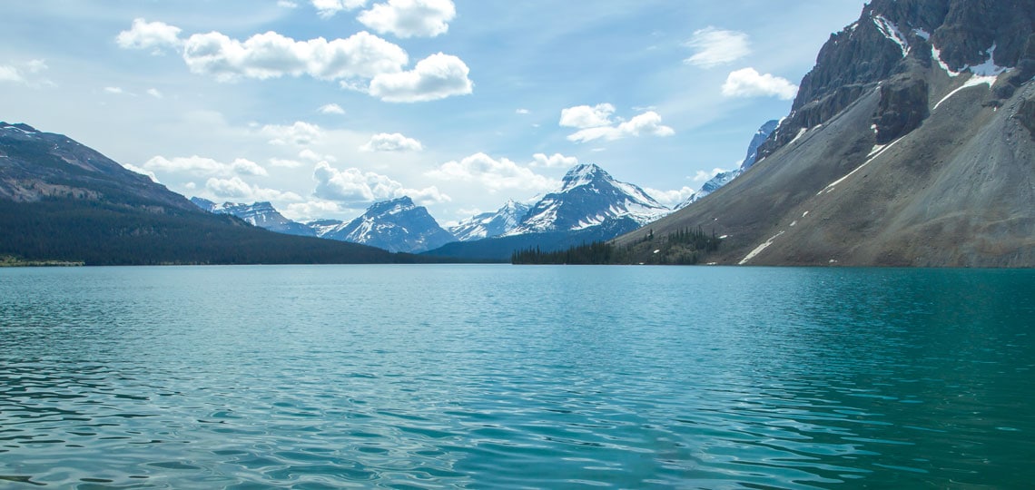 A large, still lake spans wide to distant snow capped mountains.