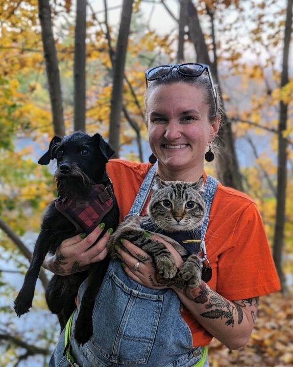 Zoryana smiles at the camera holding a small black dog and a striped cat with trees in the background.