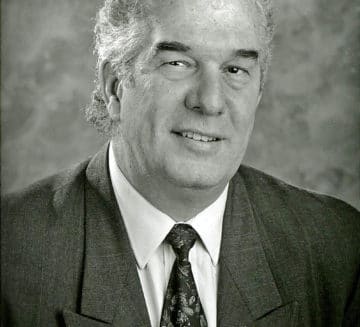A black and white photo of Peter. He wears a suit and tie and has short, curly hair.