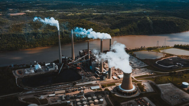 An aerial view of a large industrial plant. Smoke billows from many smoke stacks and fills the sky.