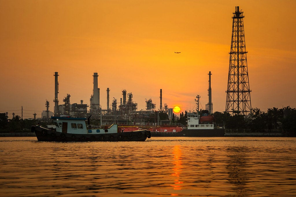 The sun rises in an orange sky behind an industrial complex on the shore of a lake.