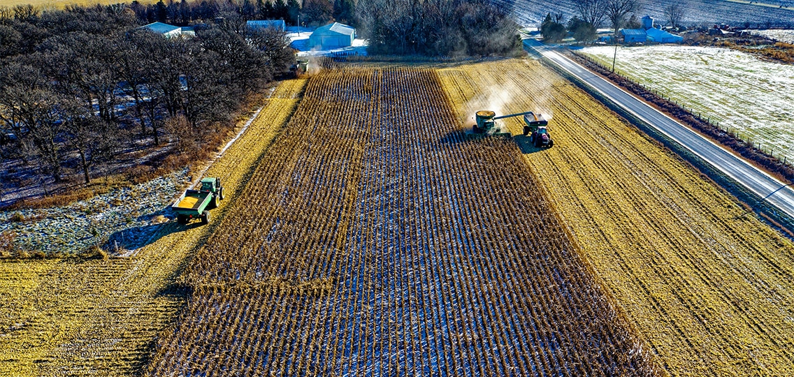 An aerial view of farmland. Two tractors harvest crops in the field.