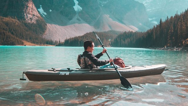 A person paddles in a kayak on clear blue water. Evergreen trees and mountains are on the distant shoreline.