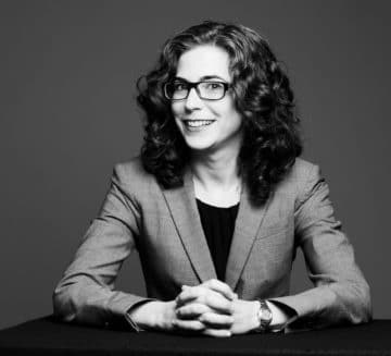 A black and white photo of Lindsay. She sits at a desk with her hands clasped together. She wears a suit jacket and square glasses. She has curly, dark hair.