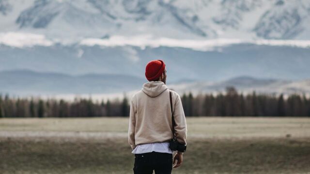 A person stands on a flat grassy plane. They look out to the trees and snow covered mountains in the distance.