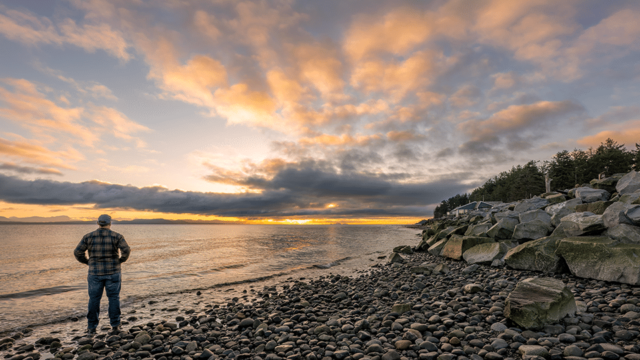 A person stands on a rocky shore looking out at the water and cloudy sunset.