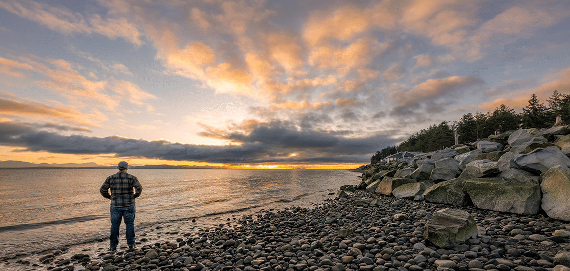 A person stands on a rocky shore looking out at the water and cloudy sunset.