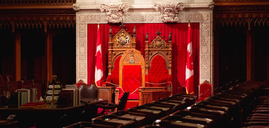 The interior of a Canadian Parliament building with large, red chairs next to Canadian flags and many empty seats for members.