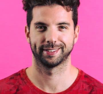 Eric smiles at the camera. He stands against a pink background and wears a red shirt. He has dark facial hair and has short, brown hair.