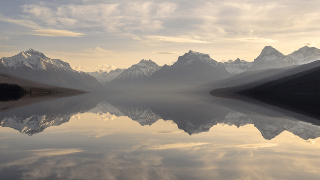 A still lake reflects distant snowcapped mountains at sunrise.