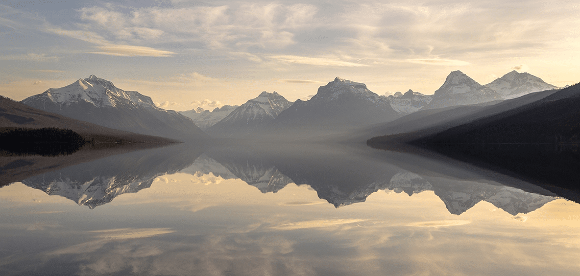 A still lake reflects distant snowcapped mountains at sunrise.