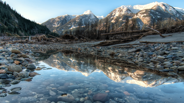 A pond of water is surrounded by a rocky shore. Fallen and bare trees stand in front of snow capped mountains in the distance.