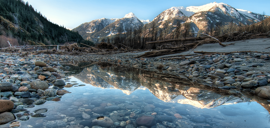 A pond of water is surrounded by a rocky shore. Fallen and bare trees stand in front of snow capped mountains in the distance.