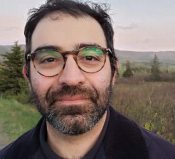 Ali stands outside in a green field. He wears round tortoiseshell glasses. He has short, brown hair and dark facial hair.