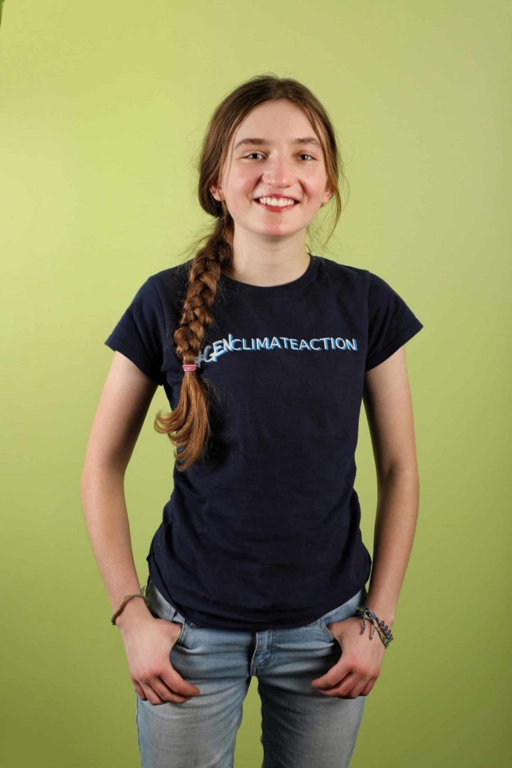 Zoe smiles and stands in front of a green background. They have a long braid and wear a hashtag gen climate action t-shirt.