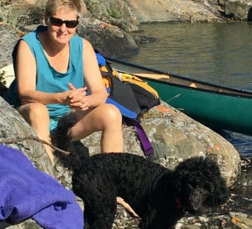 Anna sits on large rocks next to a lake. She has short, blond hair and wears sunglasses. A black dog stands at her feet and a canoe is in the water behind her.