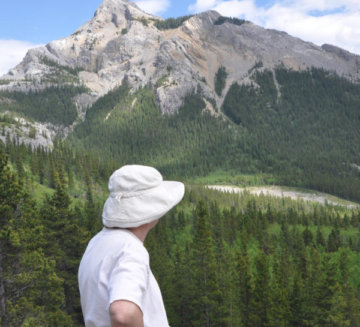 Barbara looks out at a large landscape of evergreen trees and a large mountain in the background. She wears a white shirt and hat.
