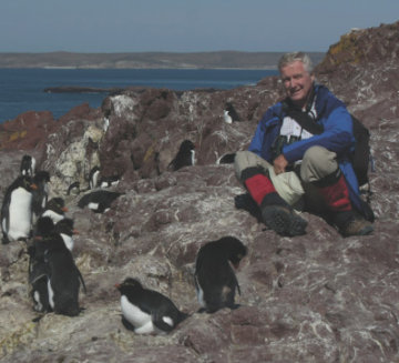 Cliff sits on the ground surrounded by penguins. He wears a blue jacket and hiking boots.