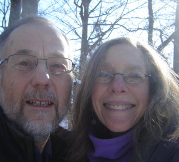 a close up of Judy and Jim. They smile while standing outside. Jim wears glasses and has white facial hair. Judy also wears glasses and has long hair.