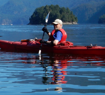Joe sits in a kayak on the calm water and holds a paddle. He wears a red life jacket and a hat.
