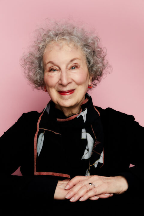 Margaret sits with her hands together. She wears a black shirt with a black and white scarf. She has short, curly grey hair.