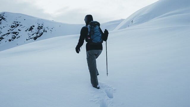 A person uses a hiking pole to walk through deep snow on mountains. They wear a dark winter coat and blue backpack.