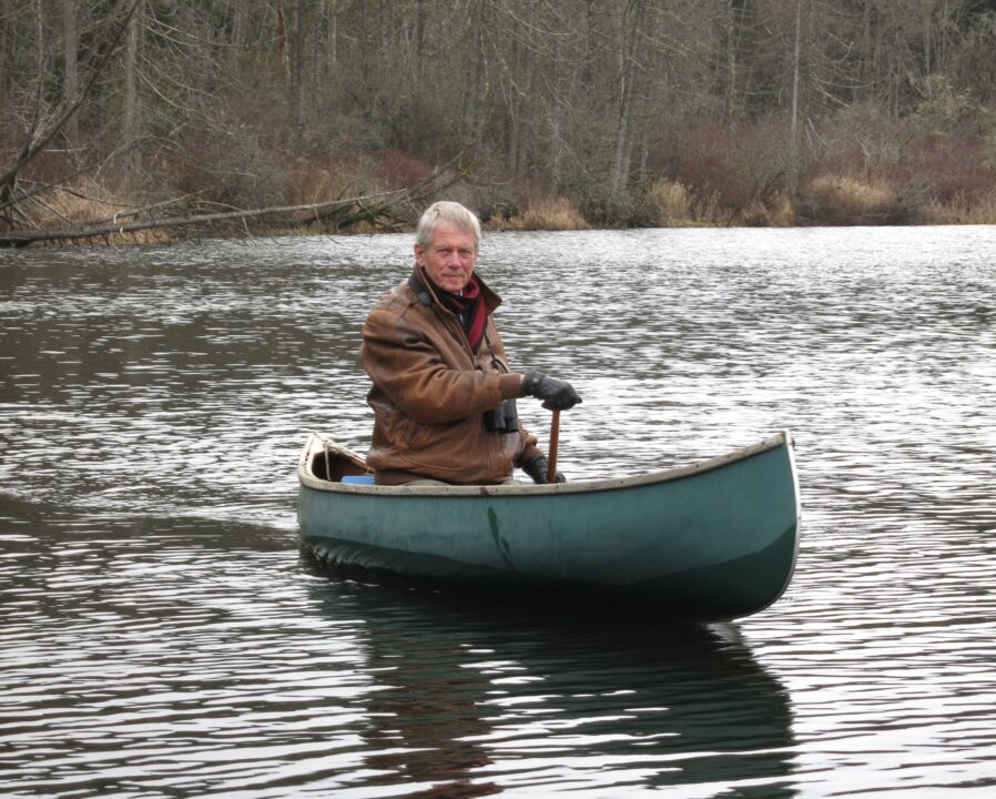 Robert sits in a canoe on the still water. He holds a paddle in the water. He has short, white hair and wears a brown jacket with gloves.