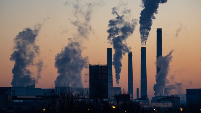 Thick smoke rises from many industrial smoke stacks at sunset. The grey smoke mixes with the orange sky.
