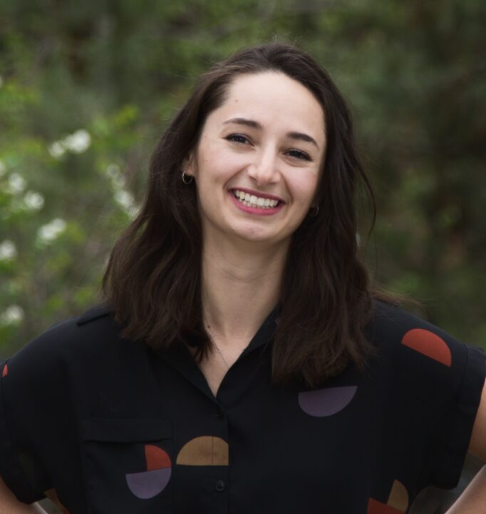 Katarina Young smiles, wearing a dark shirt with some shapes on, in front of a forest