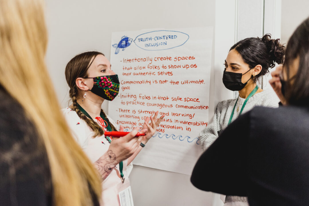 Two ladies talk to each other. One has a floral facemask on, the other has a black facemask on. Two other people are in the foreground listening. A piece of flipchart paper is attached to the wall behind them, which says Truth Centred Inclusion on it