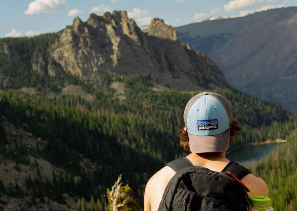 A person wearing a Patagonia cap backwards and a backpack sits with their back to the camera. They are facing a moutain and overlooking a forest and river