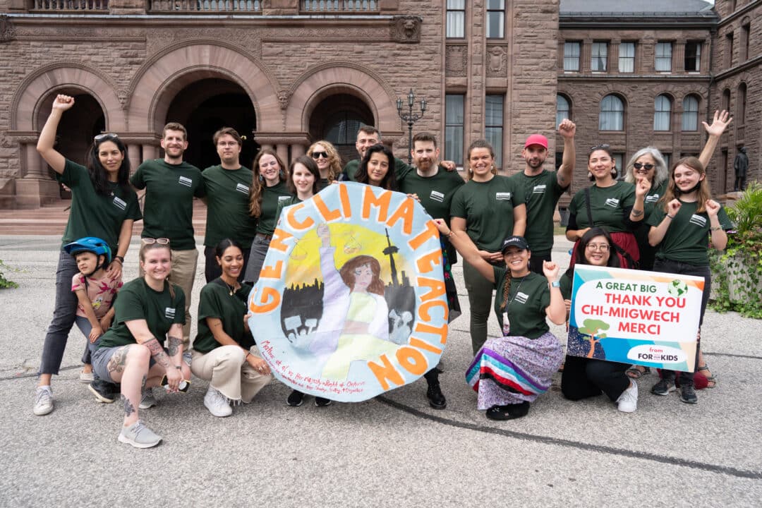A group of young people stand together holding a large round sign that says Gen Climate Action outside Queen's Park in Toronto. They are wearing dark green tshirts. Some are standing with their arms raised, some are kneeling. A second sign says 'A great big thank you'