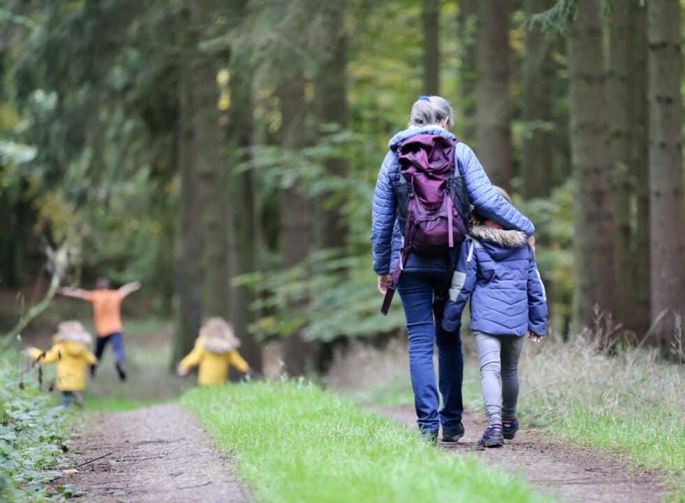 An adult and a child walk down a grassy path with tyre tracks in a forest. The adult has their arm around the shoulders of the child. Three children can be seen in the distance playing, wearing brightly coloured coats