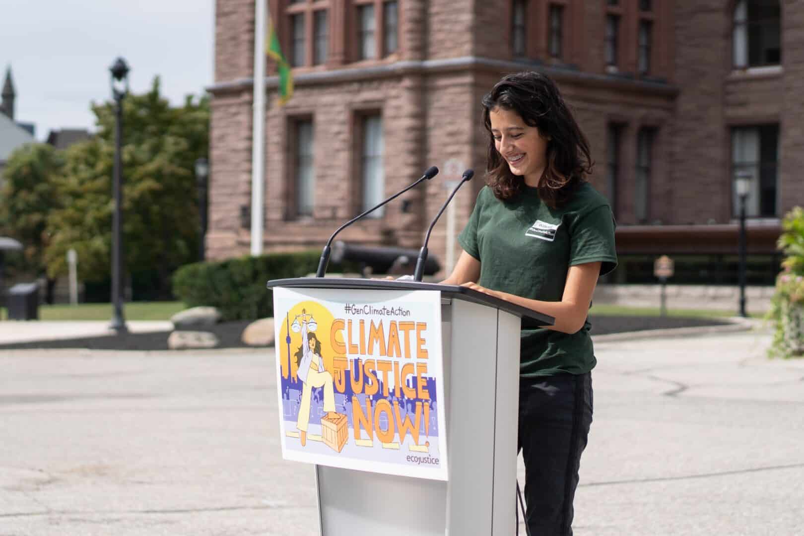 A young woman stands in front of a podium with the words Climate Justice Now on the front.