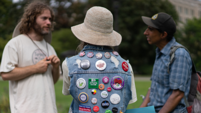 Three people at a climate rally talk to each other. One wears a denim jacket with numerous protest pins and their back towards the camera.