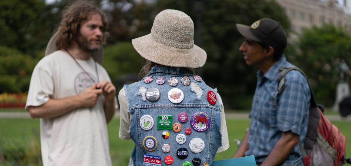 Three people at a climate rally talk to each other. One wears a denim jacket with numerous protest pins and their back towards the camera.