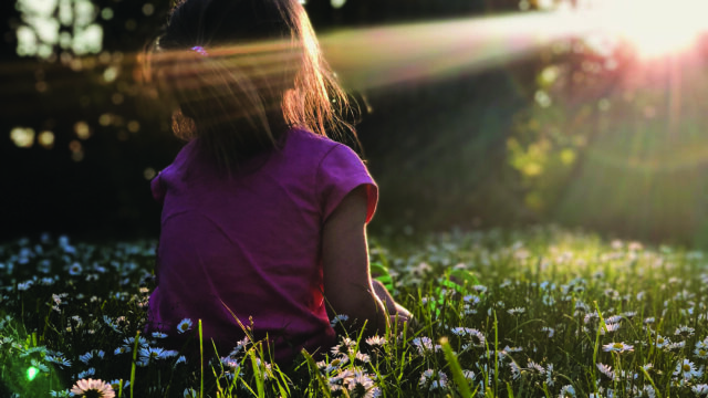 young girl sitting in a field with trees at sundown with the rays hitting her face