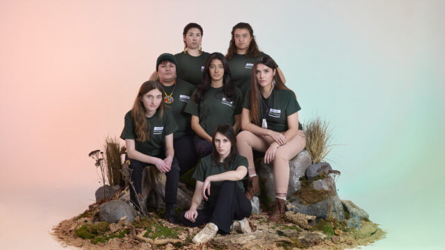 A group of youth stand together with serious expressions. They are sitting on grass and rocks brought into a studio.