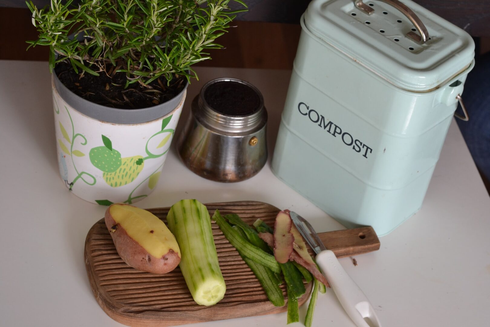 green plant on a white and purple floral ceramic pot. Also a cutting board with peeled vegetables and a compost bin.